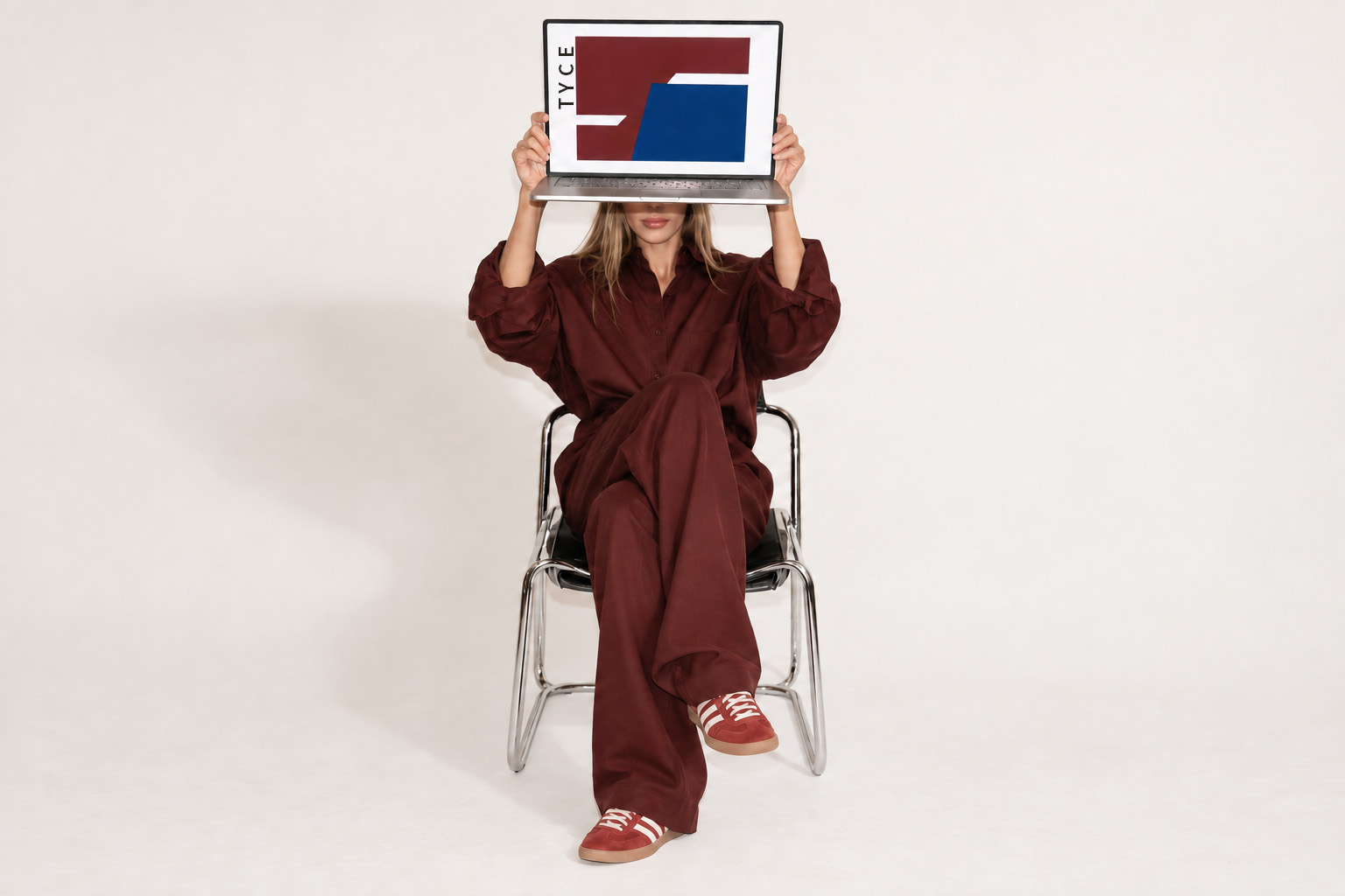 Seated woman in burgundy holding a laptop over her face, TYCE artwork on screen